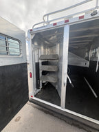 Rear tack room of horse trailer with carpeted saddle racks, multiple hooks for gear storage, aluminum walls, open rear door showing organized space for saddles and horse equipment.