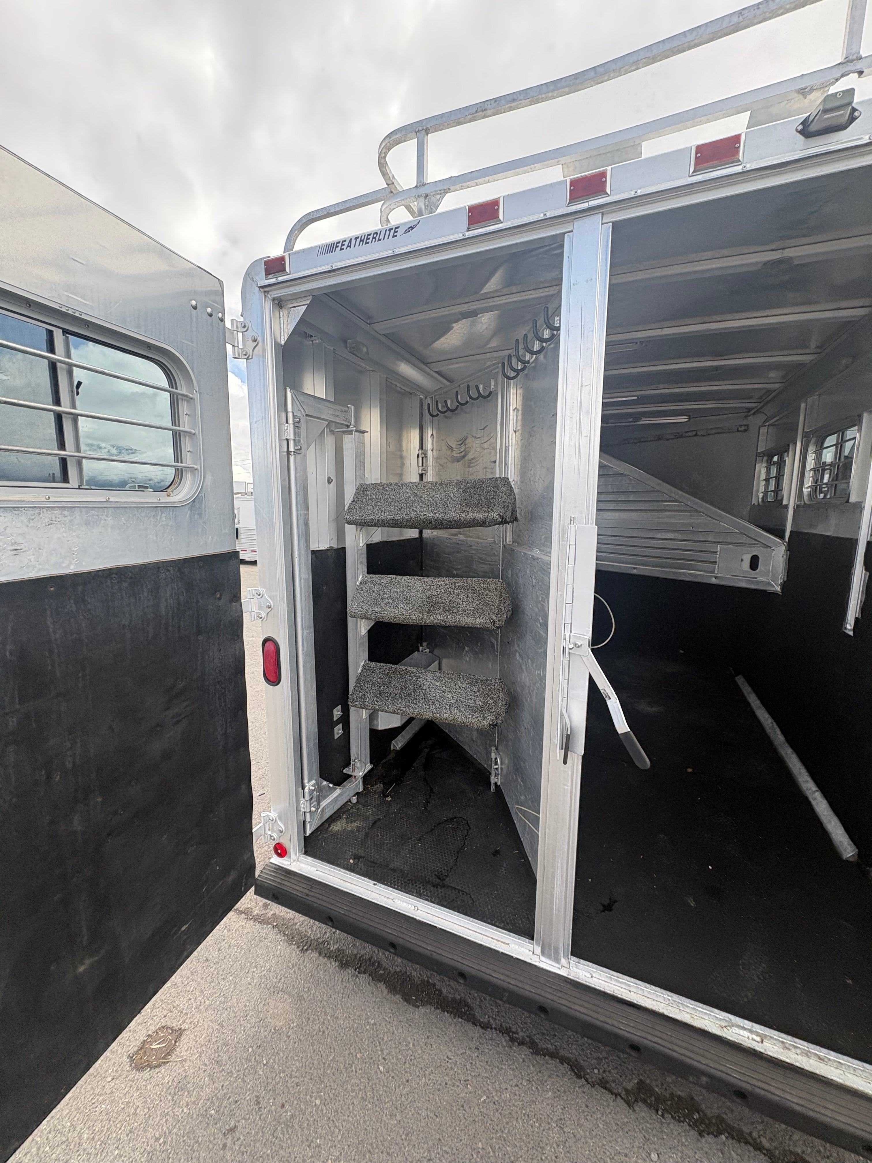 Rear tack room of horse trailer with carpeted saddle racks, multiple hooks for gear storage, aluminum walls, open rear door showing organized space for saddles and horse equipment.