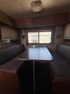 interior dining booth with cushioned bench seating, central table, side windows with blinds, and warm wood cabinetry inside horse trailer living quarters