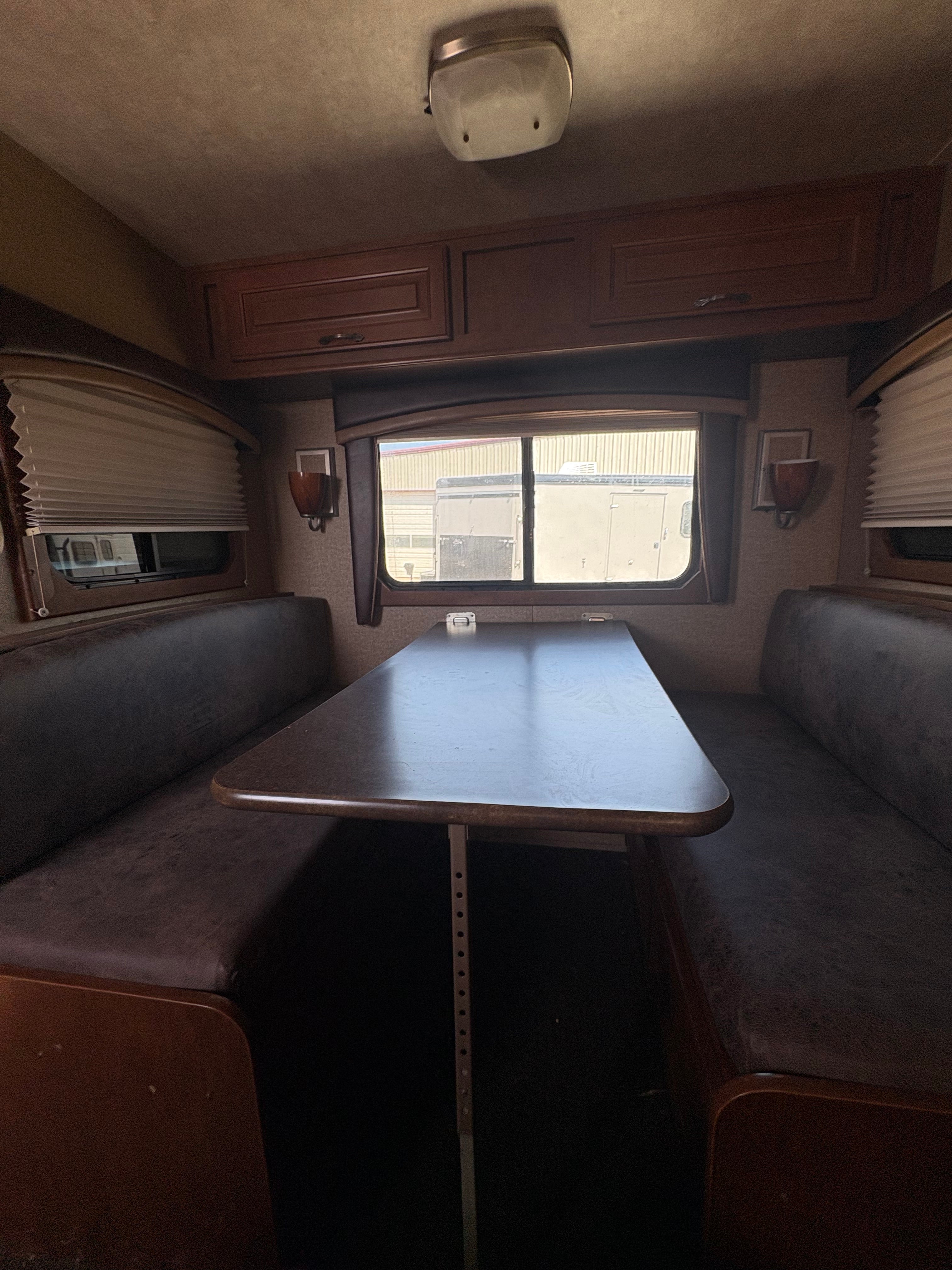 interior dining booth with cushioned bench seating, central table, side windows with blinds, and warm wood cabinetry inside horse trailer living quarters