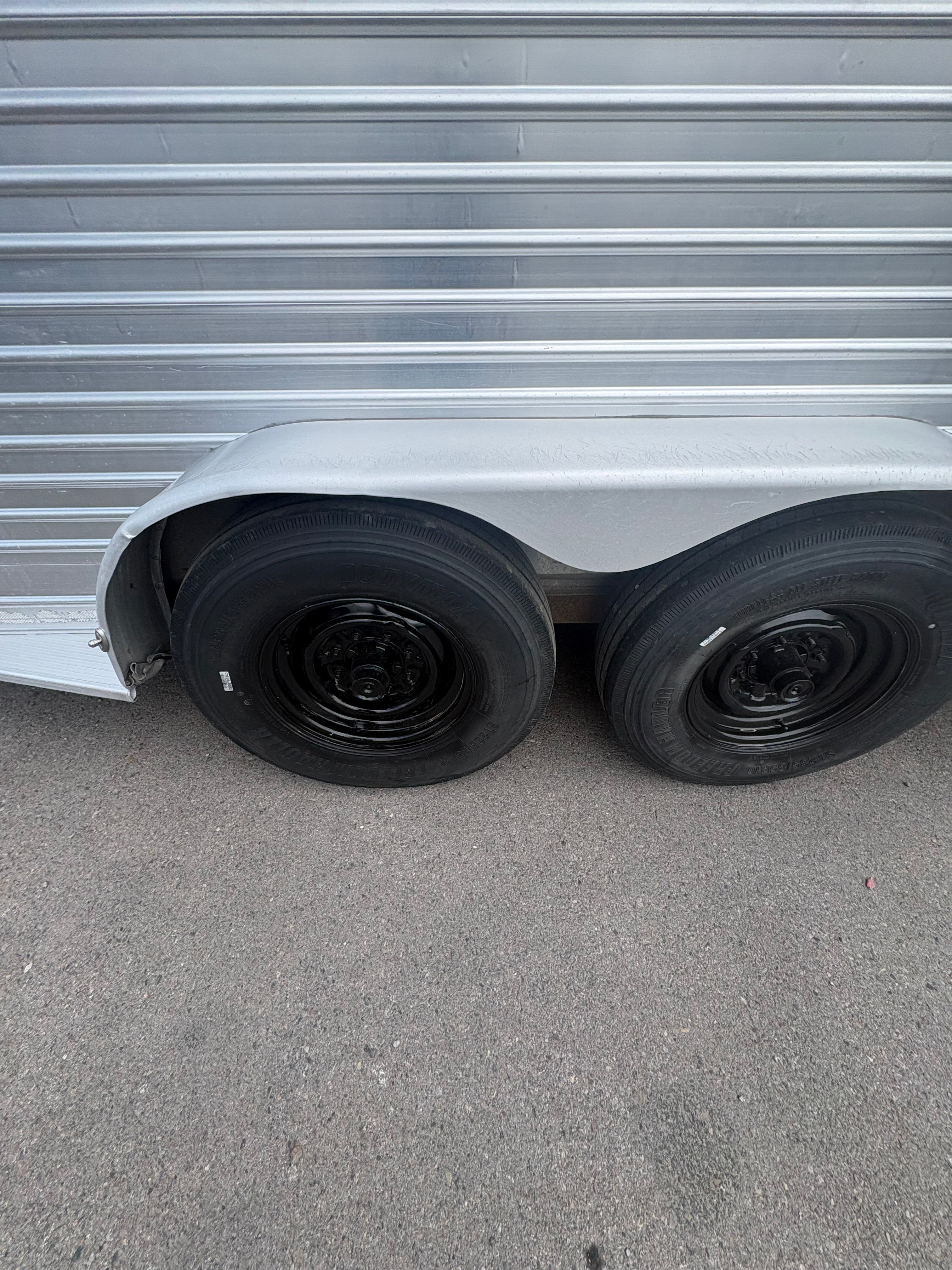 Close up of dual axle horse trailer wheels with black steel rims and heavy duty tires, aluminum fender above tires, textured metal siding detail visible on livestock trailer exterior.