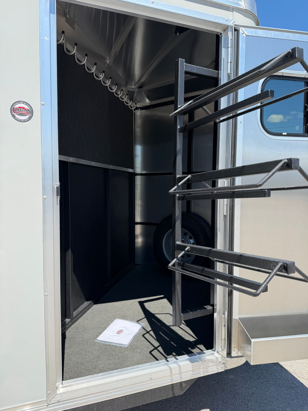 heavy duty swing out saddle rack system inside Logan Coach Crossfire trailer with multiple padded saddle holders, upper bridle hooks, black carpeted walls, and spacious tack storage area.
