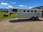 Logan Coach Crossfire gooseneck 4 horse trailer parked beside fenced pasture with mountain backdrop, featuring gray aluminum exterior, four horse windows, tandem axles, and spacious front sleeping area under bright blue sky