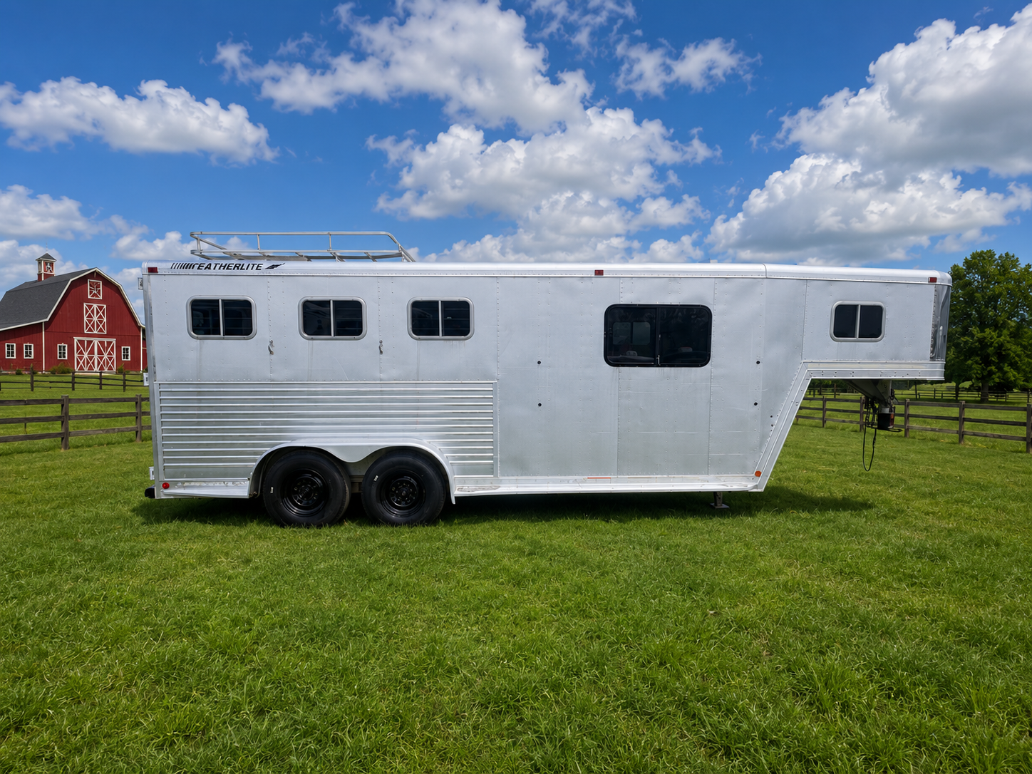 Aluminum gooseneck horse trailer parked on green grass under blue sky, dual axle design with black wheels, smooth metal exterior, roof rack on top, long body livestock trailer used for hauling multiple horses.