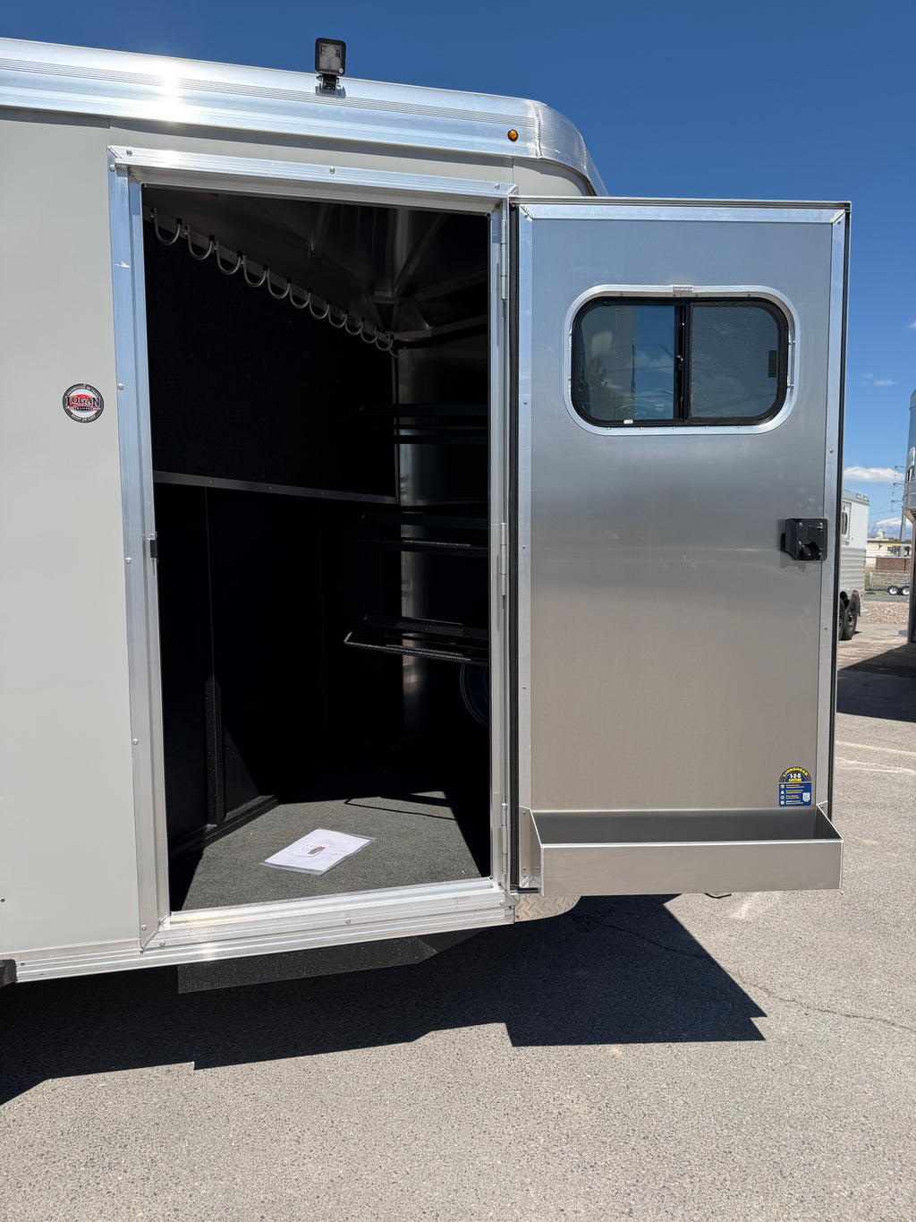 open front tack room on Logan Coach Crossfire horse trailer featuring carpeted interior walls, bridle hooks, saddle storage space, aluminum tack door, and organized horse gear compartment.