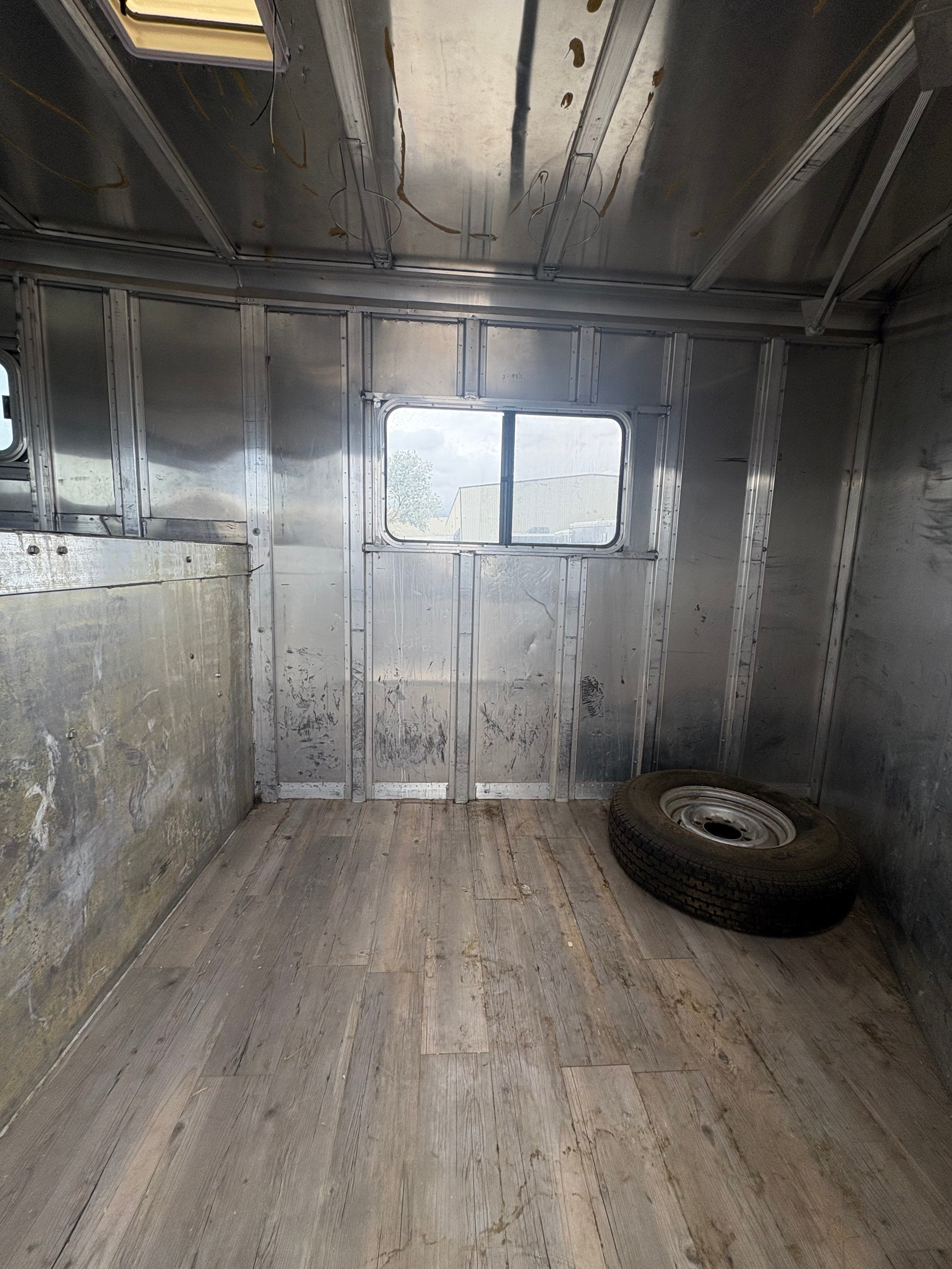 Aluminum horse trailer storage compartment with wood plank flooring, spare tire placed on floor, metal walls with scuff marks, enclosed utility space for equipment or tack storage.