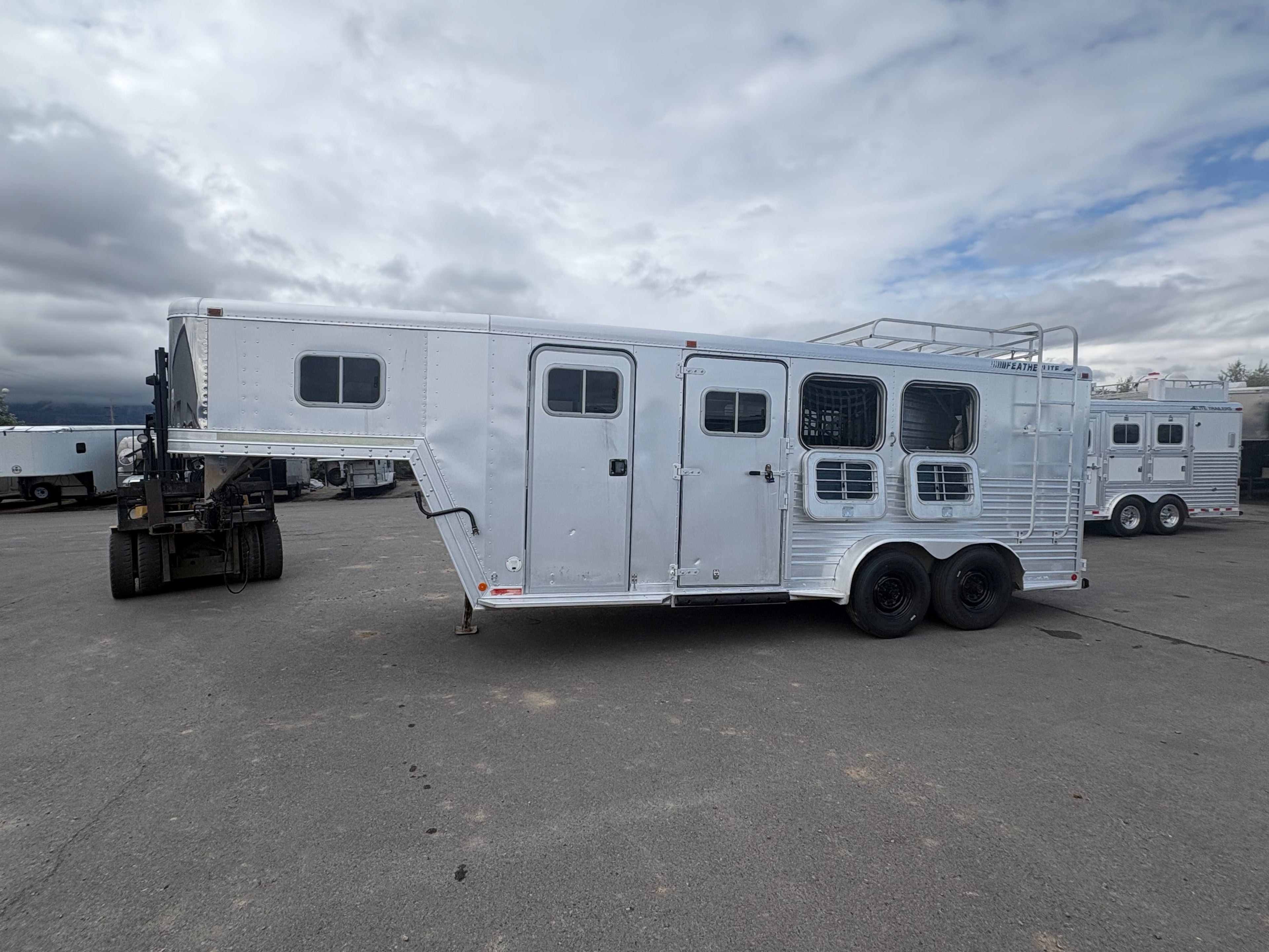 Aluminum gooseneck horse trailer exterior showing side access door, multiple horse windows with bars, dual axle wheels, riveted metal panels, parked on paved lot with cloudy sky background.