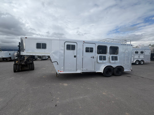 Aluminum gooseneck horse trailer exterior showing side access door, multiple horse windows with bars, dual axle wheels, riveted metal panels, parked on paved lot with cloudy sky background.